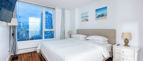 A modern hotel room with a large bed, a bedside table, a TV mounted on the wall, and a window offering a city view.