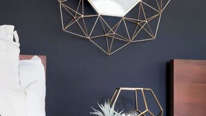 Una habitación con un espejo dorado de marco geométrico, una cama blanca con una colcha blanco y negro, una mesita de noche de madera con una planta en maceta y una pared oscura.