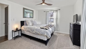 Une chambre à coucher comprenant un lit, un ventilateur de plafond, une table de chevet avec une lampe, une commode et une fenêtre avec des rideaux.