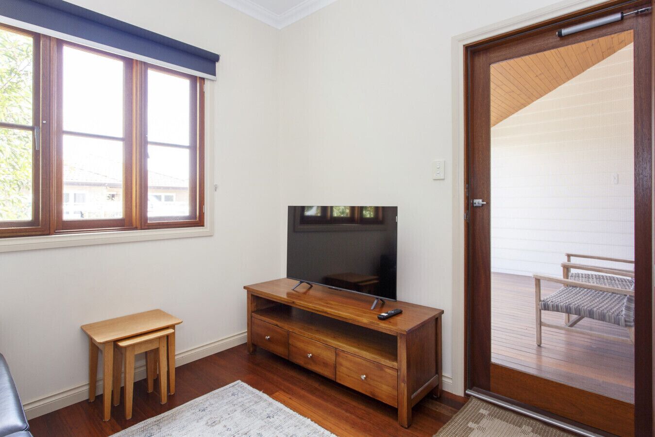 Une pièce comprenant un meuble de rangement en bois pour appareils multimédias, un téléviseur, une petite table en bois et une porte donnant sur un espace extérieur avec un banc.