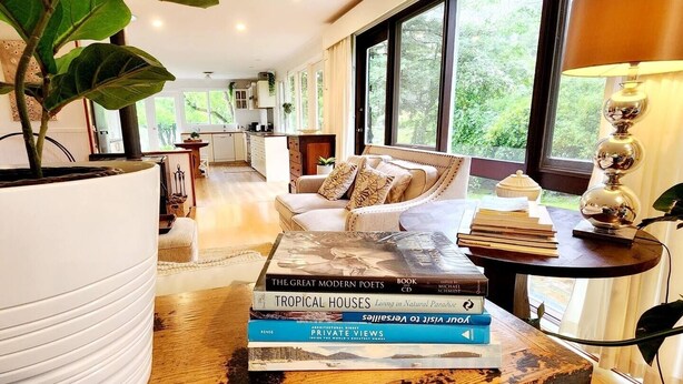 Un salon moderne avec un canapé, une table basse sur laquelle sont posés des livres, une grande plante et une vue sur l’extérieur grâce à de grandes fenêtres.