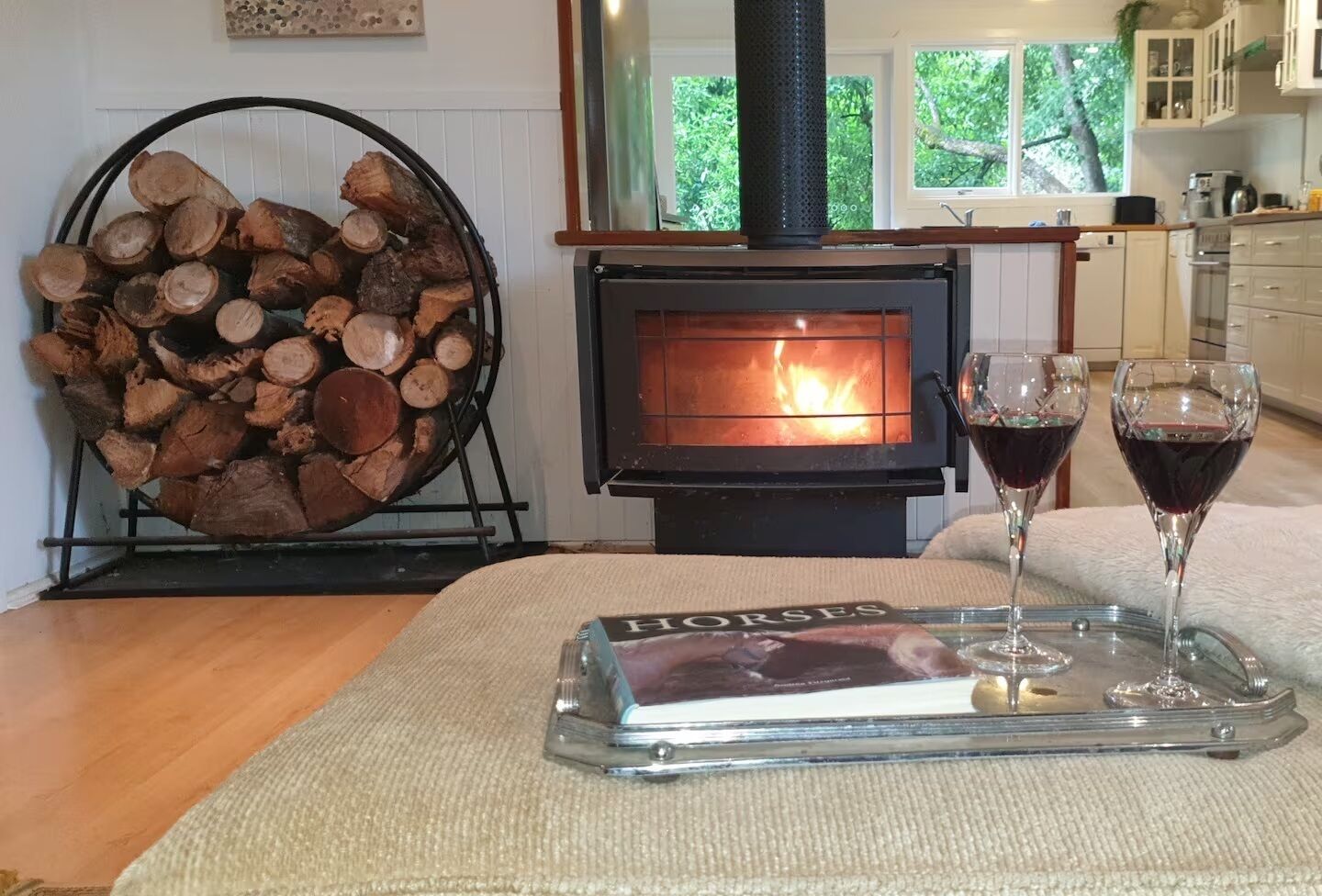 Een knus vertrek met een houtkachel, een stapel hout en twee glazen rode wijn op een dienblad.