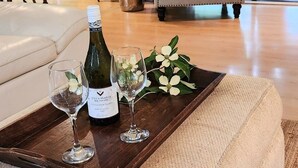 Un salon chaleureux avec une table basse en bois sur laquelle reposent une bouteille de vin et des verres, un canapé beige et une vue sur la verdure grâce à de grandes fenêtres.