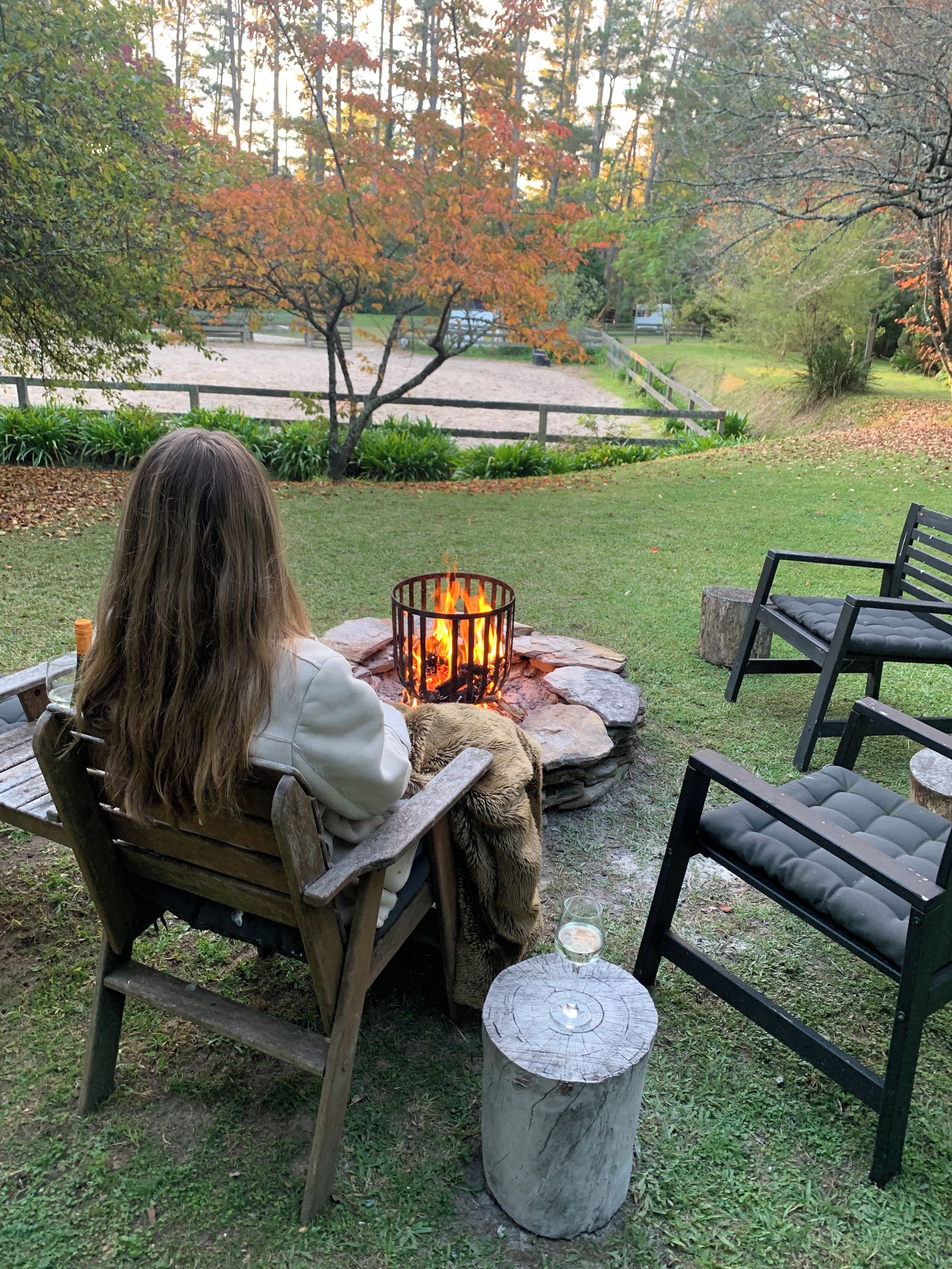 A person sitting by a fire pit in a grassy outdoor area with a wooden bench and chairs, surrounded by trees and a wooden fence.