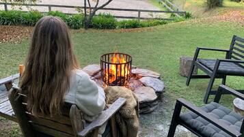 A person sitting by a fire pit in a grassy outdoor area with a wooden bench and chairs, surrounded by trees and a wooden fence.