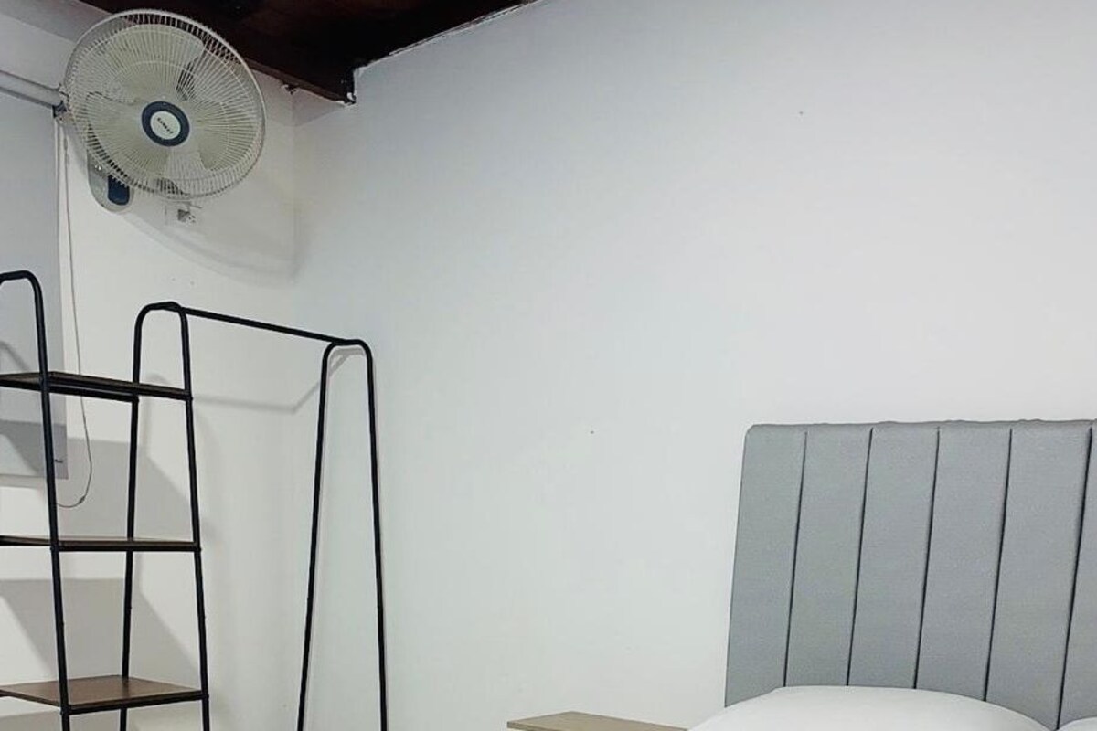 Een slaapkamer met een bed, nachtkastje, een ventilator aan de muur en een metalen schapunit.