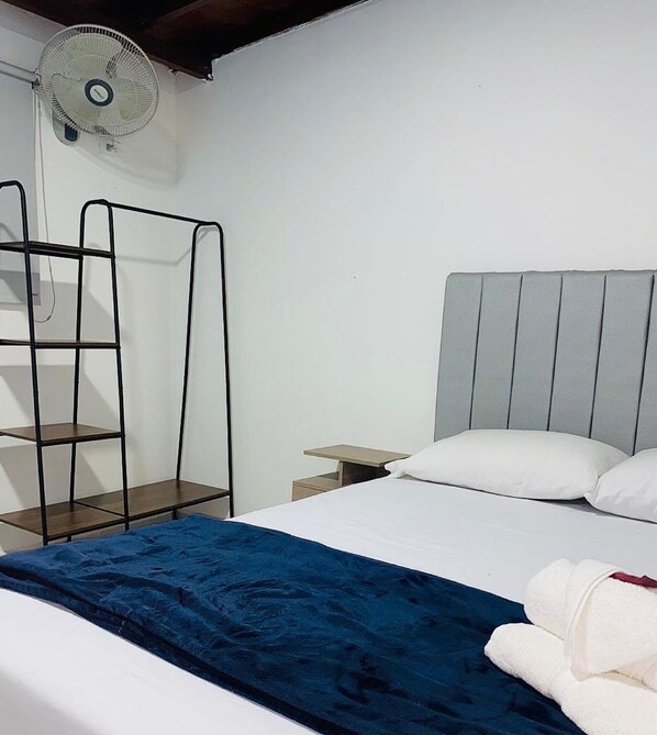 A bedroom with a bed, a bedside table, a wall-mounted fan, and a metal shelf unit. - HOTEL SAN JUAN 70 EXPRESS  (Medellín)