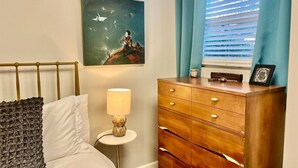 Une chambre à coucher avec un lit, une commode, une lampe, un tableau et une fenêtre avec des rideaux.