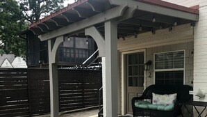 A covered patio area with a pergola, a black outdoor chair, and a white building in the background.