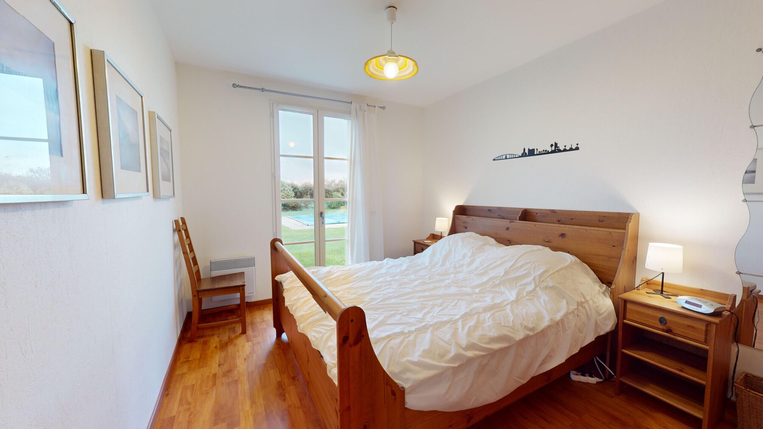 Ein Schlafzimmer mit einem hölzernen Bett, einem Nachttisch mit Lampe, einem Sessel und einem Fenster mit Vorhängen.