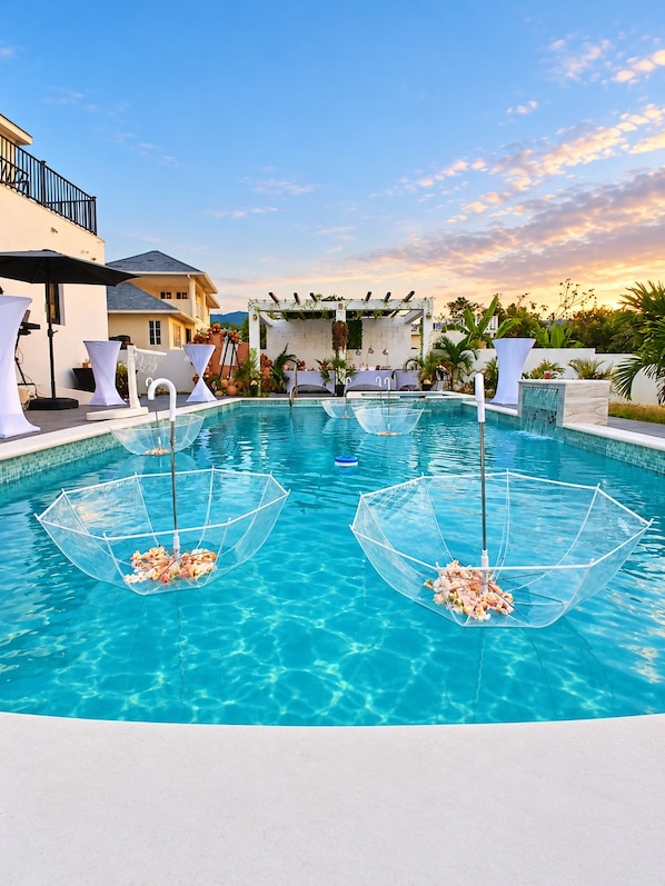 Una piscina azul cristalina con dos sombrillas transparentes llenas de flores, rodeada de mobiliario blanco y un pérgola blanca con follaje.