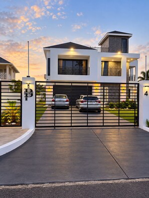 A modern two-story house with a gated driveway, two cars parked inside, and a well-maintained lawn. - Modern Oceanview Villa. All Bedrooms with Private Bathrooms. Private Pool (St. Ann Parish)