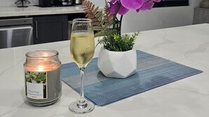 Ett kök med mörka skåp, en vit bänkskiva, ett ljus med etiketten ”Royal Sage”, ett glas vitt vin och en krukväxt med lila blommor.