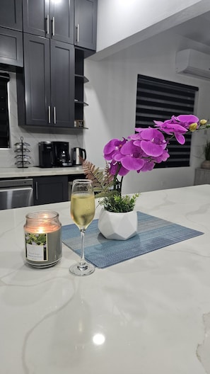 Eine Küche mit dunklen Schränken, einer weißen Arbeitsplatte, einer Kerze mit der Aufschrift 'Royal Sage', einem Glas Weißwein und einem Topf mit violetten Blüten.