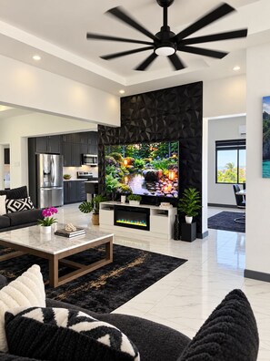 A modern living room with a flat-screen TV, a ceiling fan, and a kitchen in the background.