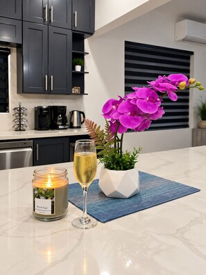 Una cocina moderna con armarios oscuros, una cafetera, una vela y una copa de vino blanco sobre una encimera de mármol.