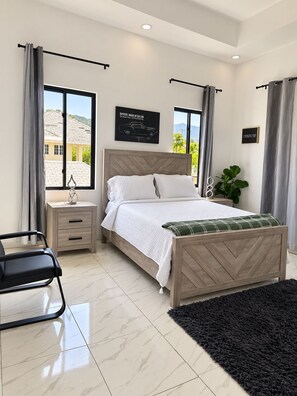 Un dormitorio con cama, mesitas de noche, una silla, un ventanal con cortinas y vistas a una casa.