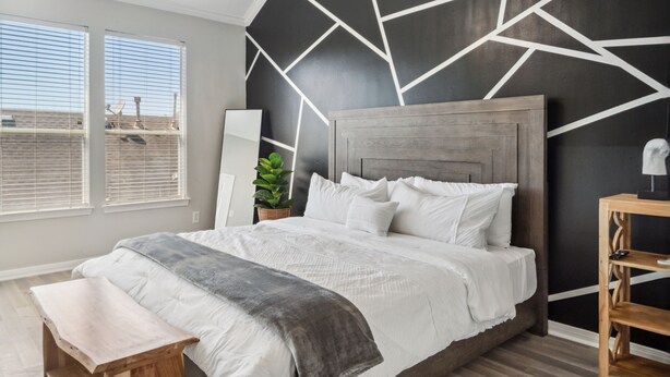 A modern bedroom with a geometric wall design, a wooden headboard, a large bed with white bedding, a bench, and a wooden nightstand. - 5 Minutes to Downtown + GARAGE! Near Med Center/NRG/George R Brown Convention (Houston)