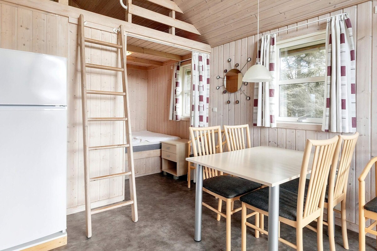 A cabin interior with a bunk bed, a dining table with chairs, a small kitchen area, and a window with curtains.