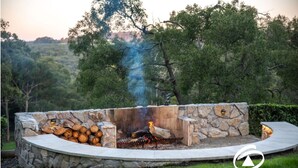 A stone fire pit with logs and a fire, surrounded by a stone wall and a wooden deck. - 'Linden Rise'  cosy farmhouse, peace & charm with views  for miles. (Pakenham Upper)