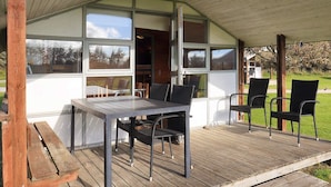 A covered outdoor seating area with a table and chairs, large windows, and a wooden deck. - Pretty Cabin in Camping Resort (Lokken)