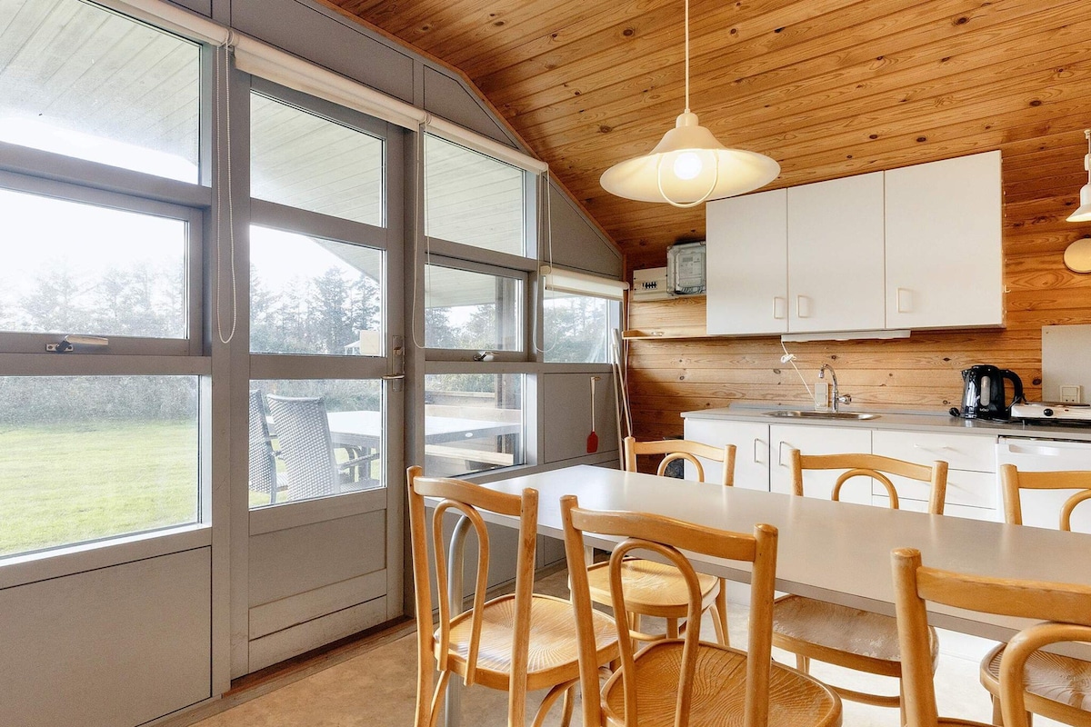 A modern kitchen with large windows, wooden ceiling, and a dining area with wooden chairs.