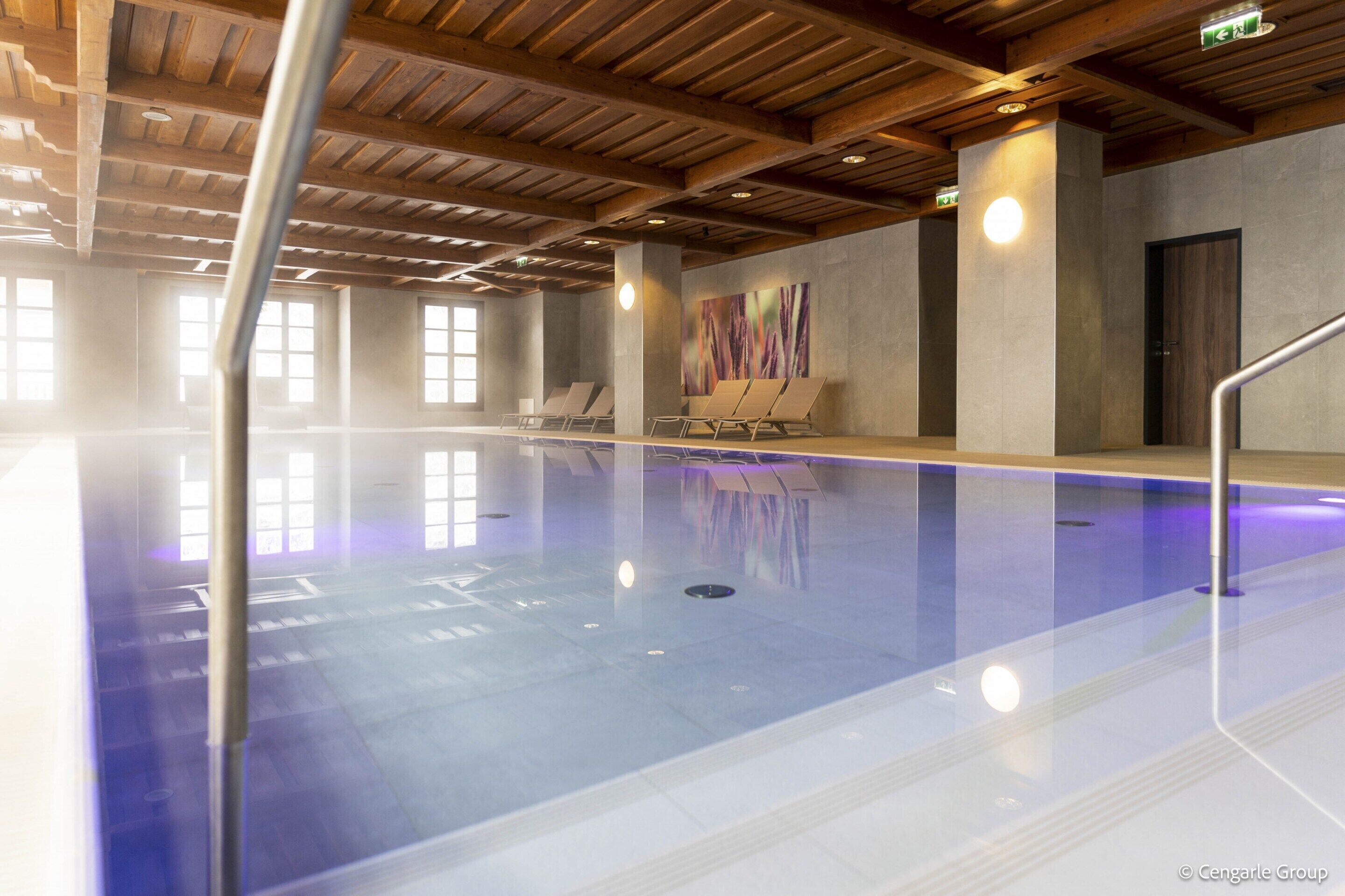A swimming pool with a wooden ceiling and walls, a few lounge chairs, and a large abstract painting on the wall.