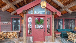 Une cabane en rondins avec une porte rouge, une couronne de Noël et un paillasson sur lequel est écrit « MAISON ».