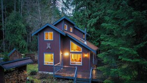 A two-story cabin with a steep roof, large windows, and a wooden deck. - Cozy Mt. Baker Escape • Fireplace • Pets (Glacier)