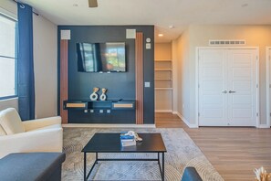 A modern living room with a sofa, a coffee table, a flat-screen TV mounted on the wall, and a white door. - ROVE LIVING/KL - Alta North Central (Phoenix)
