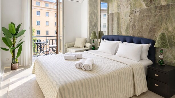 A hotel room with a bed, a nightstand, a lamp, a potted plant, and a view of the outside balcony.