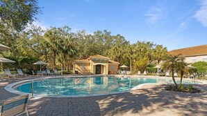 A pool area with lounge chairs and umbrellas, surrounded by palm trees and a building with a tiled roof. - Family House near Disney - 5109 (Kissimmee)