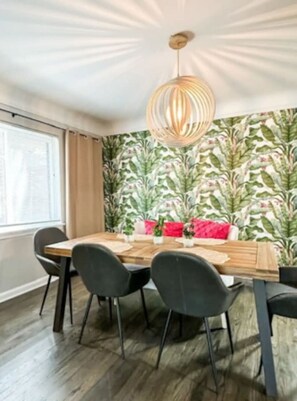 A dining area with a wooden table, black chairs, a hanging light fixture, and a wall with a leafy pattern.
