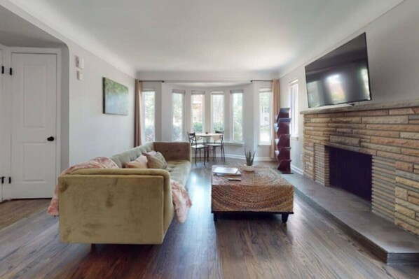 A living room with a sofa, coffee table, fireplace, TV, and large windows. - Grosse Pointe Retreat | 3BR, Sleeps 8 & Relaxing (Grosse Pointe)