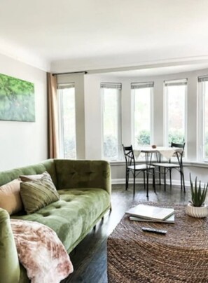 A room with a green sofa, a dining table with chairs, and a large window with curtains.