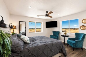 Un dormitorio con cama, dos sillas, televisor y ventanas amplias con vistas.