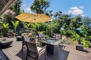 Relax on the serene outdoor deck, surrounded by lush greenery and trees, with a comfortable seating area and central fire feature.