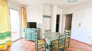 Een eetgedeelte met een tafel en stoelen, een televisie op een kast, en een deur naar een andere kamer.