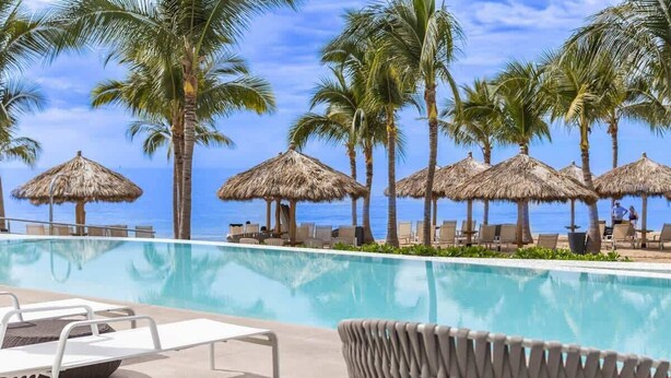A poolside area with lounge chairs, thatched umbrellas, and palm trees. - Marítima Beach Condo, 2BR, Deluxe, 2 Pools, BBQ, Balconies & Great Amenities (Flamingos)