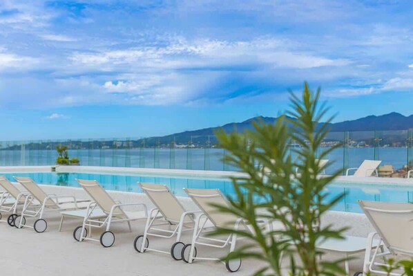 A poolside area with lounge chairs, a clear view of the sea, and distant mountains.