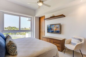 Un dormitorio con una cama grande, una silla, un escritorio, un cuadro y una ventana con vistas a zonas verdes.