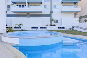 A swimming pool with a tiled edge, surrounded by a white building with balconies and potted plants.