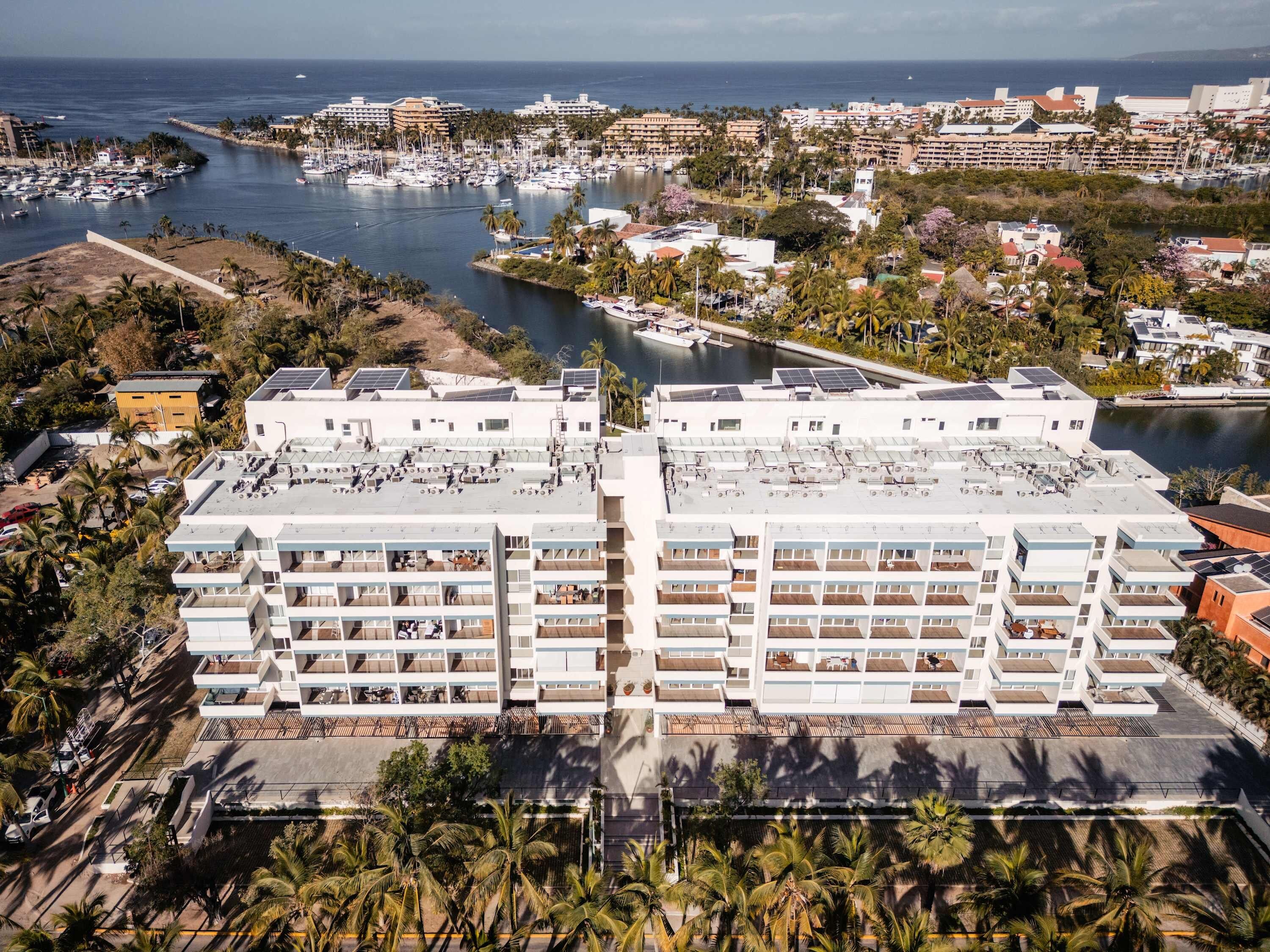 Un hotel moderno con varios pisos, paneles solares en la azotea y un puerto deportivo al fondo.