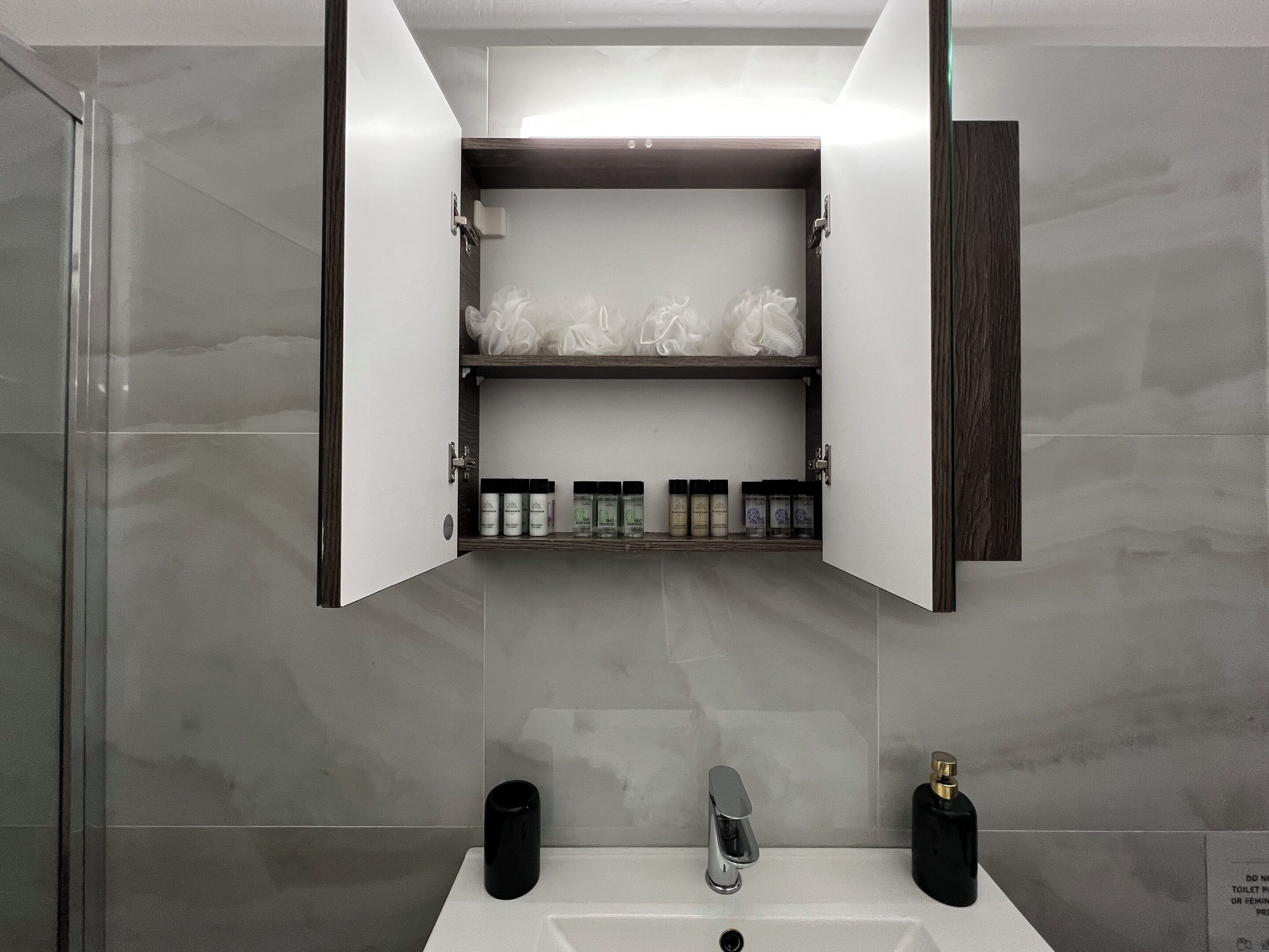 A bathroom with a white sink, a silver faucet, and a black soap dispenser. There is a mirror above the sink and a cabinet below it containing toiletries.