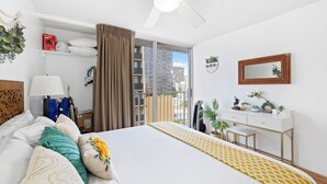 A hotel room with a bed, a desk, a chair, a ceiling fan, a window with a view of buildings, and a potted plant. - NEW! Sweet&Cozy 9thFl  1-BR king Bed (Honolulu)