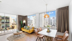 A modern living room with a yellow sofa, dining table, and chairs, large windows with curtains, and a city view. - NEW! Sweet&Cozy 9thFl  1-BR king Bed (Honolulu)