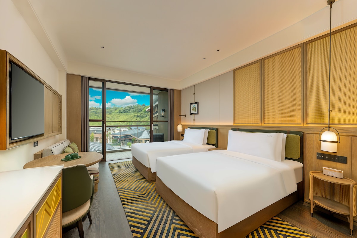 Habitación de hotel con dos camas, un escritorio, un televisor y un balcón con vista a zonas verdes.