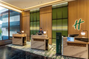 Zona de recepción de hotel con tres mostradores, un recepcionista y un letrero luminoso con la letra 'H'.
