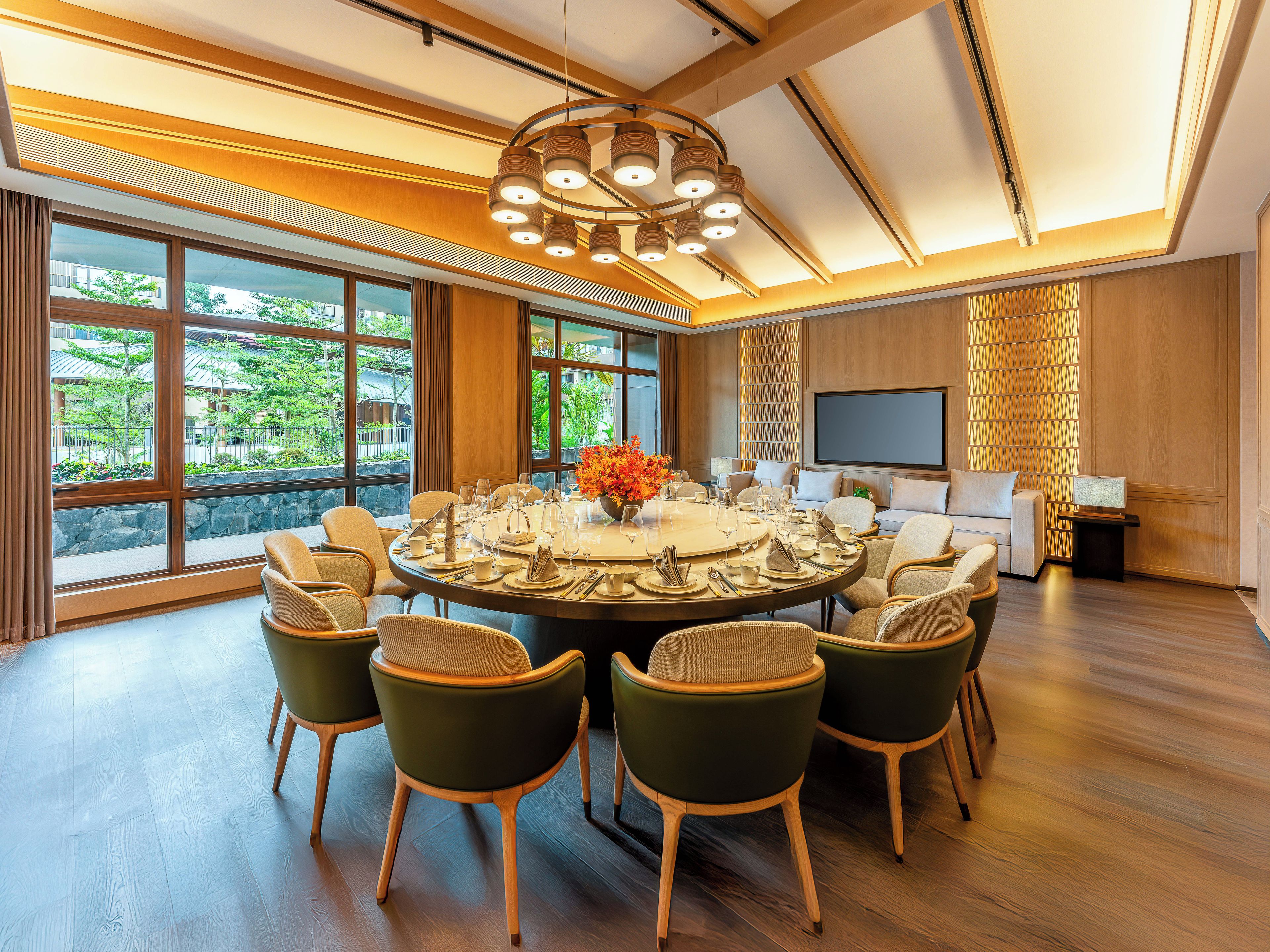 A conference room with a round table, multiple chairs, a large window with a view of greenery, and a television on the wall.