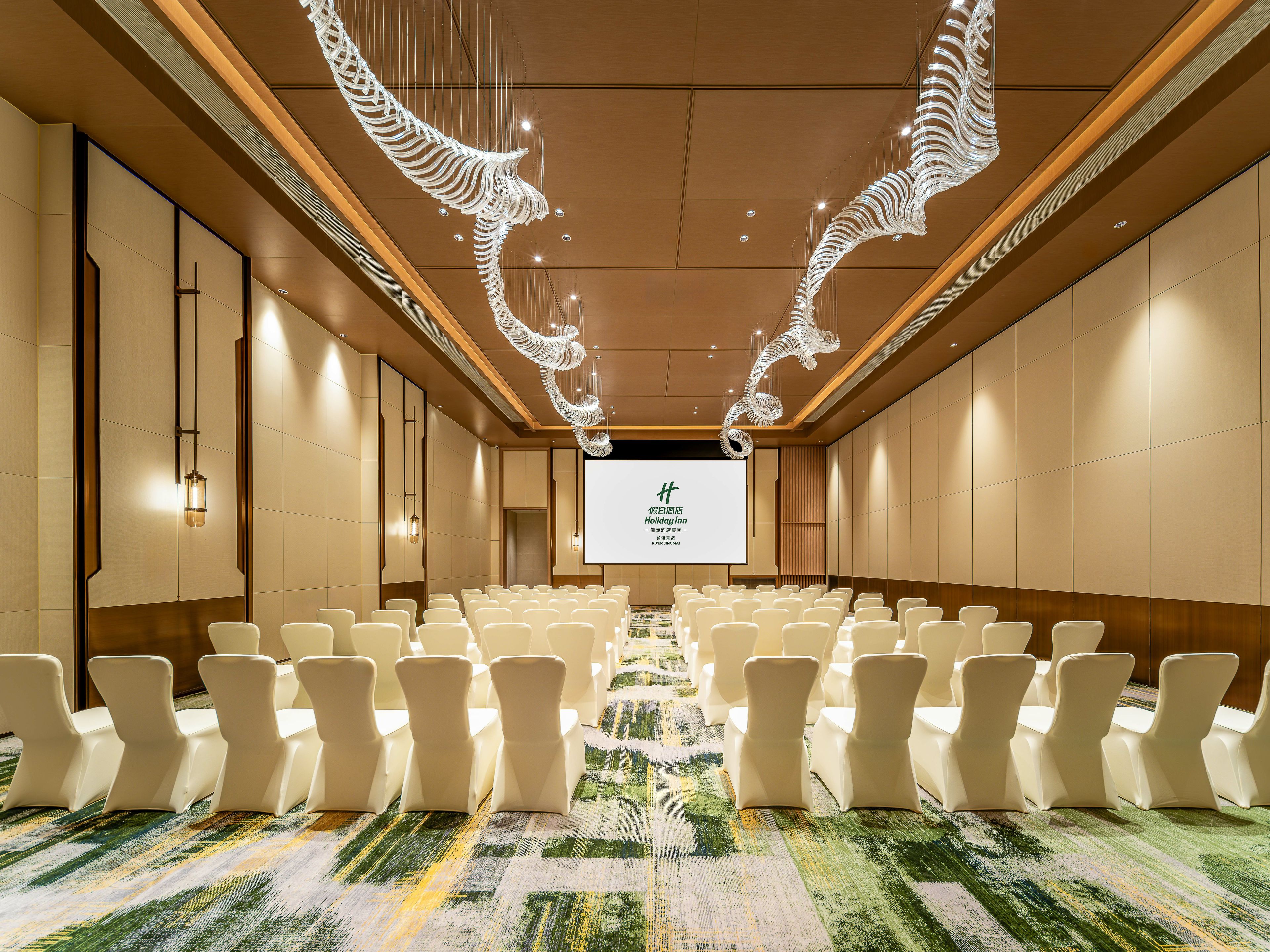 Un salón de conferencias con filas de sillas blancas, una pantalla grande al frente y una lámpara decorativa en el techo.